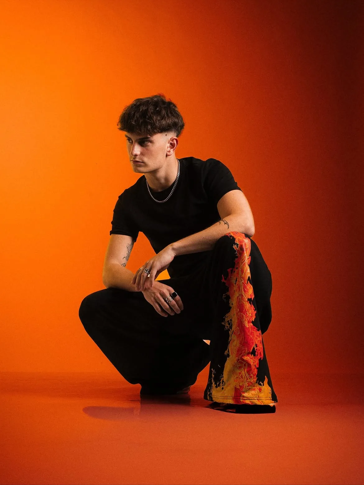 Mylan Westers crouching in black clothes and flame-print trousers against an orange studio backdrop