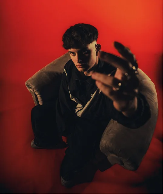 Mylan Westers seated against a red backdrop, reaching one hand towards the camera